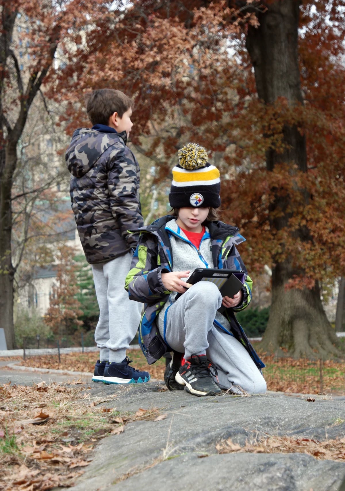 Two lower school students in Central Park, fall trees behind them. One is looking into the park and the other is crouched down looking at an ipad.