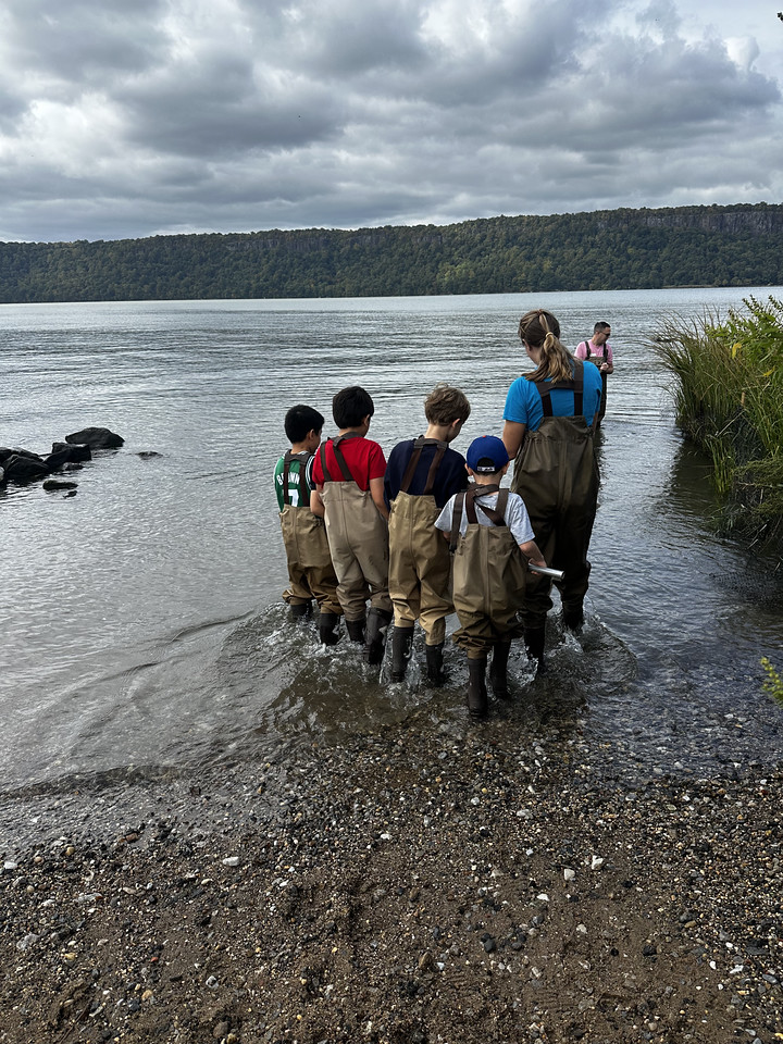 Ethical Culture students in waders collecting samples at the river’s edge with their teacher
