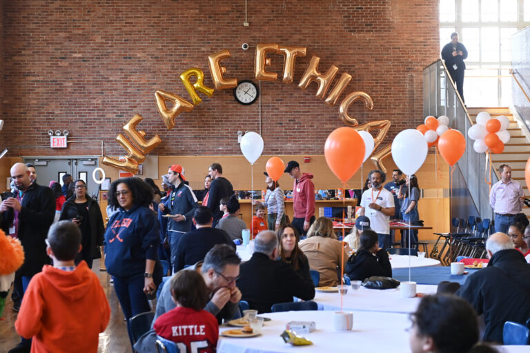 A large group of people sits and stands in the Ethical Culture Fieldston School dining room, surrounded by orange and white balloons and with