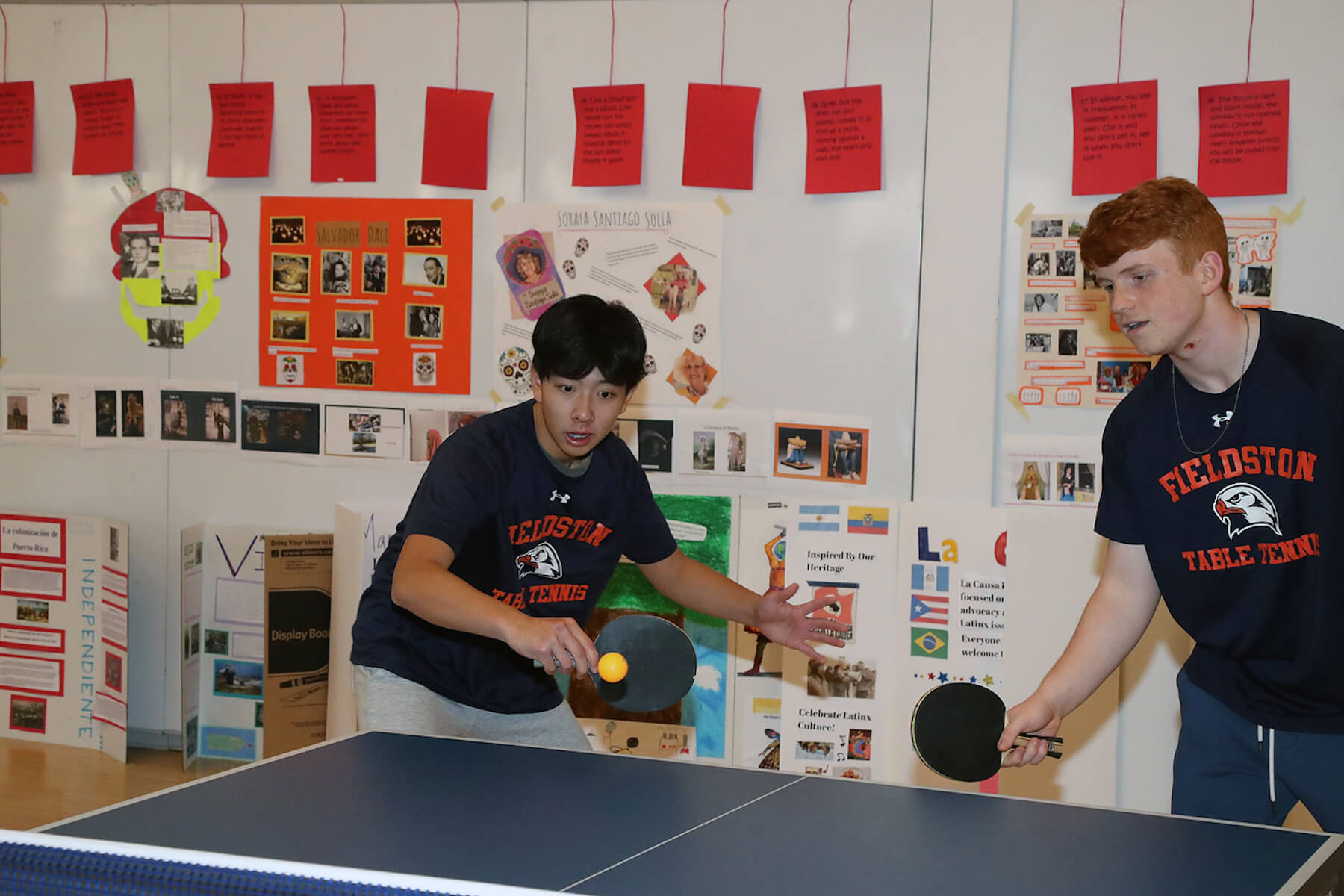 Two Fieldston Lower table tennis players compete in match. One student hits a yellow/orange ping pong ball while the other looks on, paddle raised.