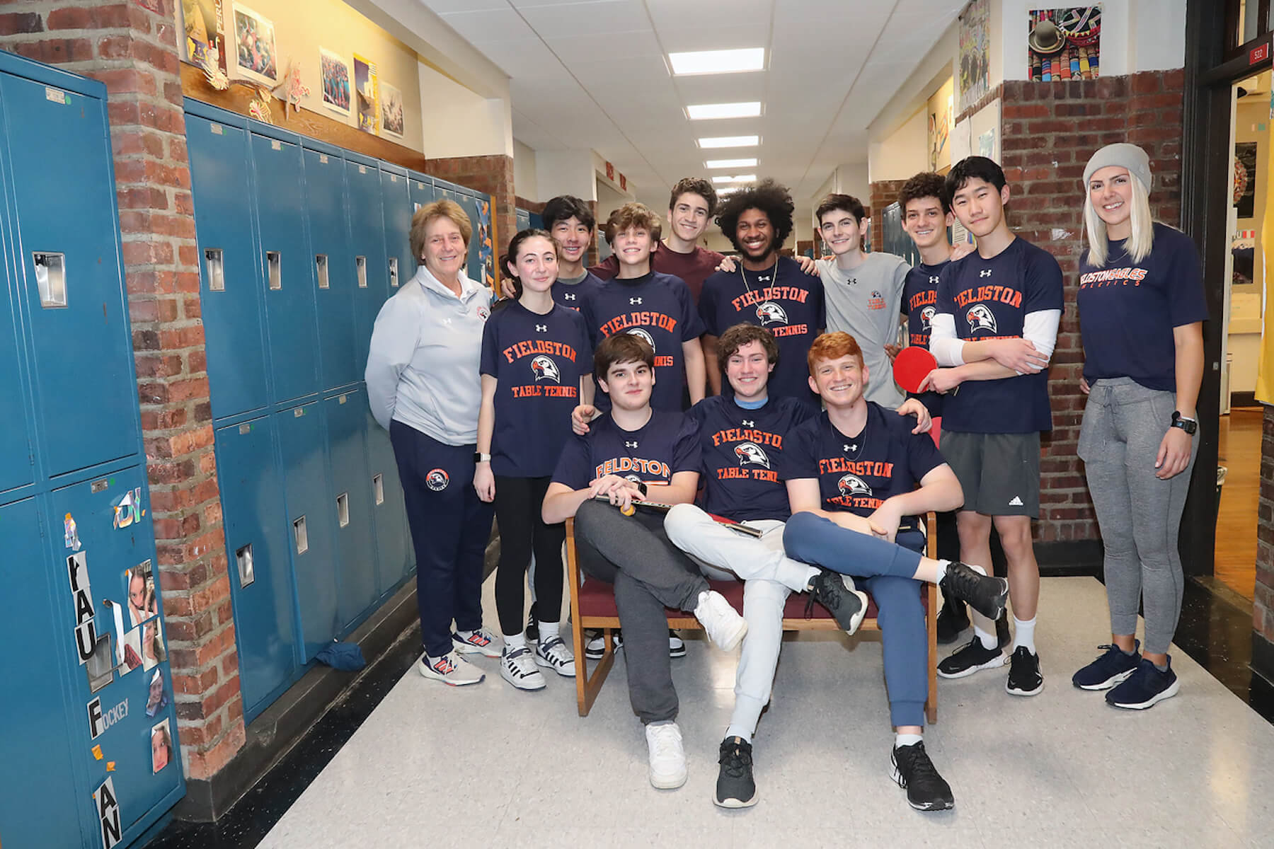 Fieldston Upper table tennis team poses for group photo in hallway; some students stand, while others sit in front on a bench.