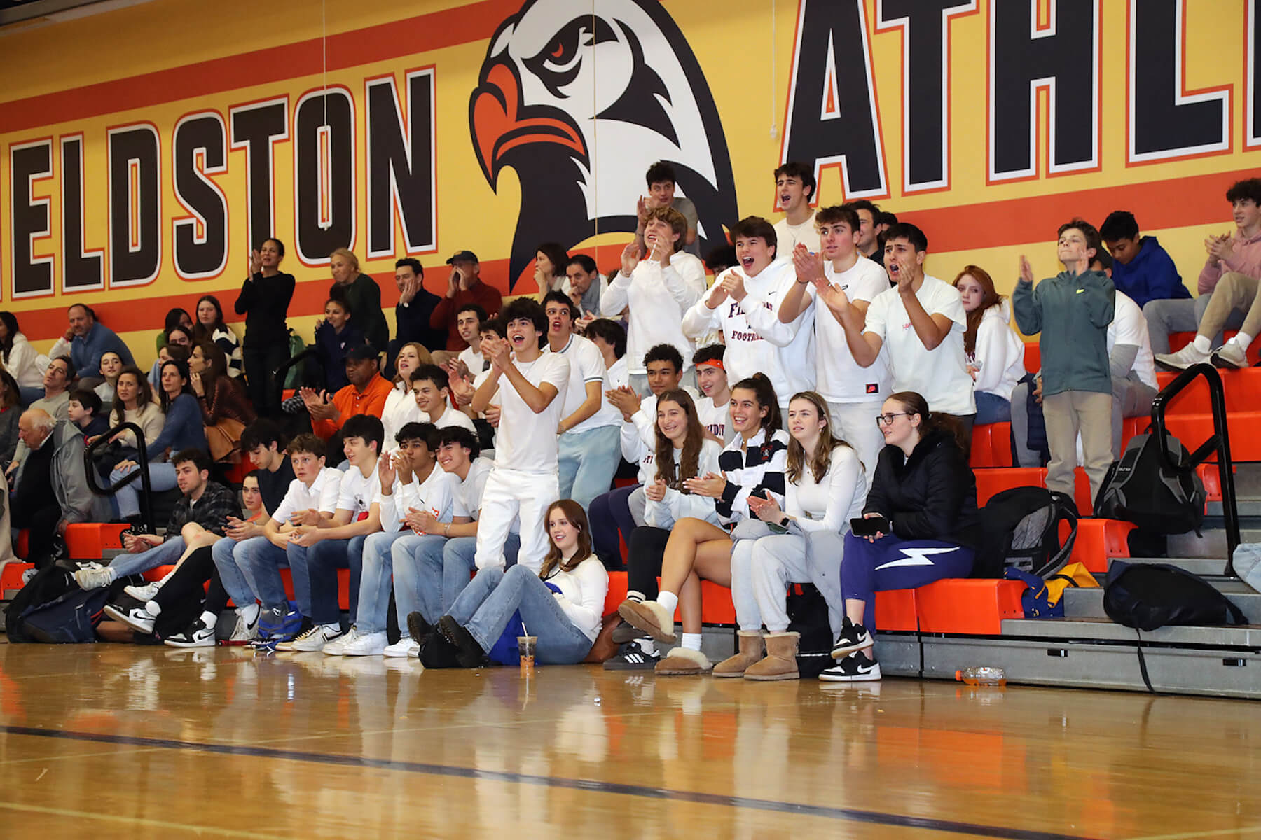 Fieldston students, wearing mostly white, cheer and smile in the bleachers as they cheer on student athletes at Winterfest.