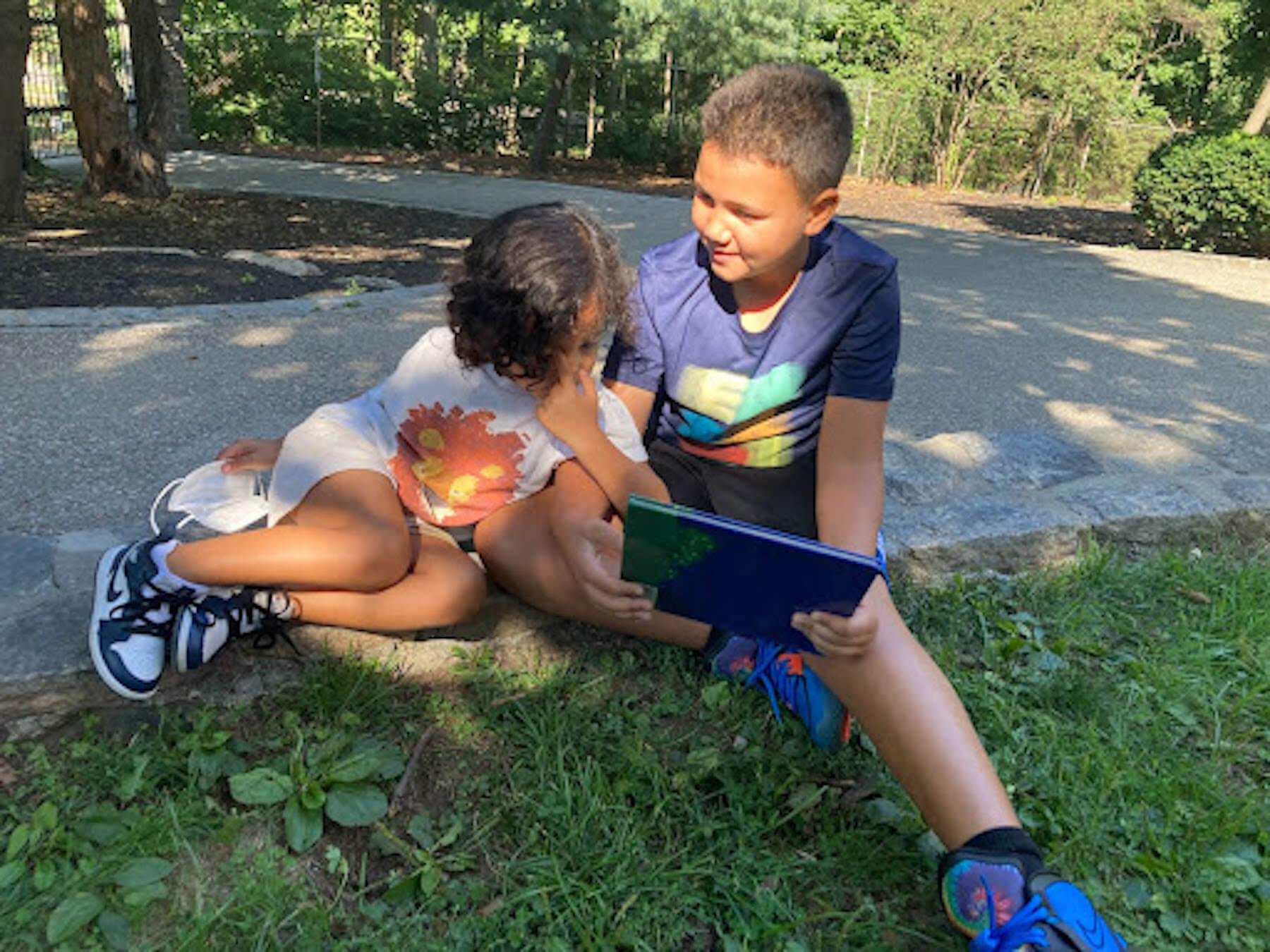 Two Fieldston Lower buddies read a book together while sitting in the grass. 
