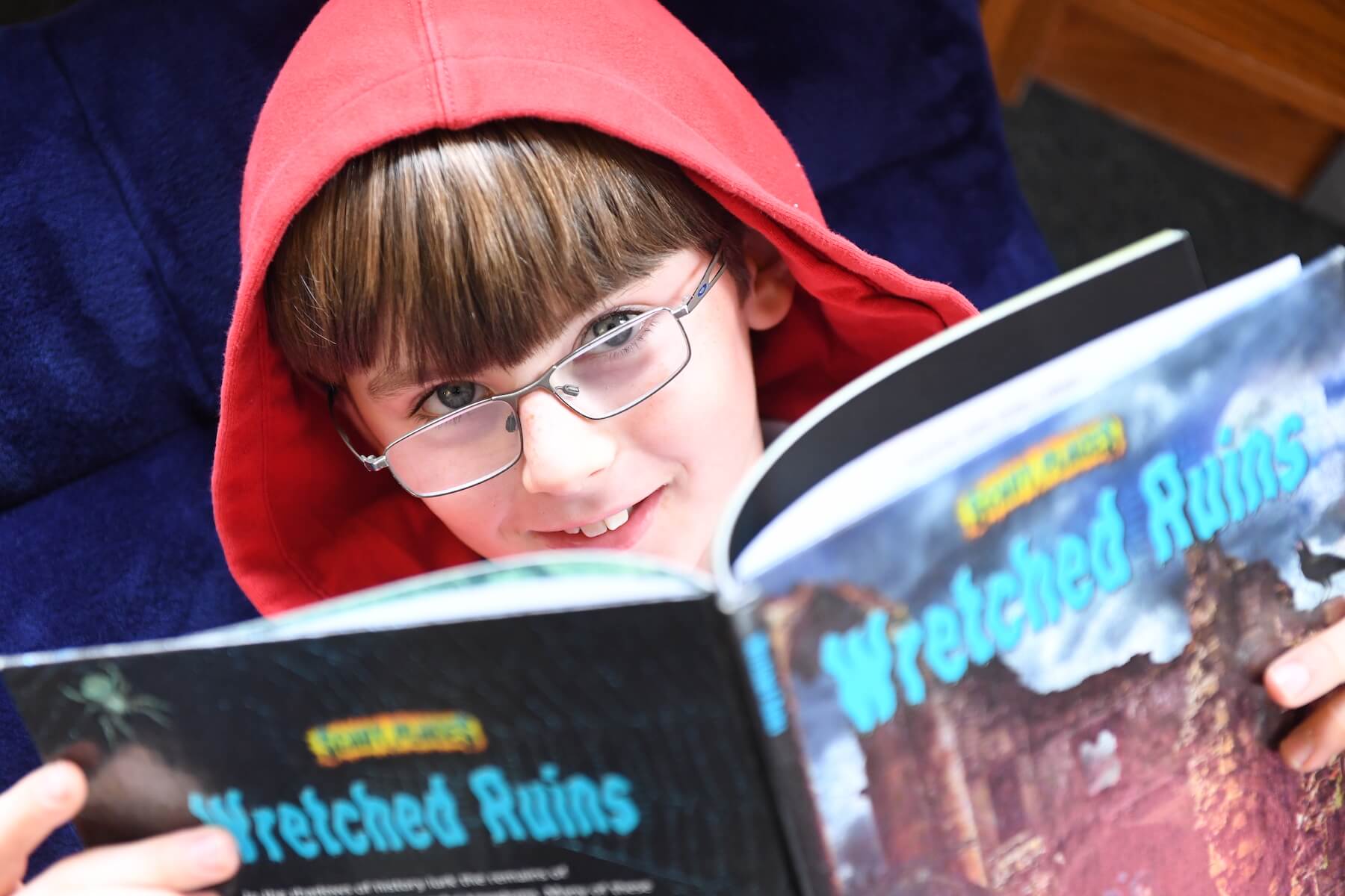 Ethical Culture Fieldston School Fieldston Lower student smiles at camera while reading a book in the library