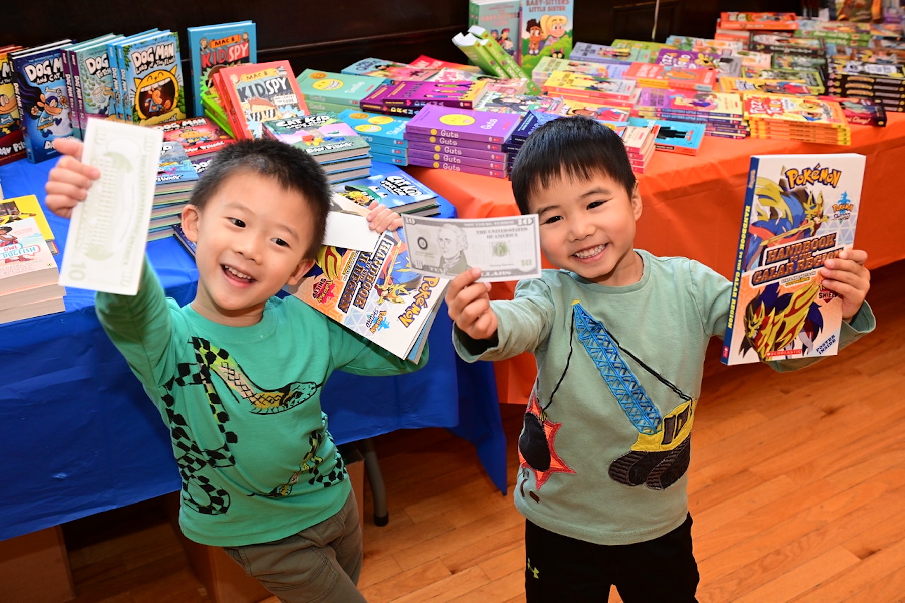 Two beaming students hold up their books and fake money at the Ethical Culture Book Fair.
