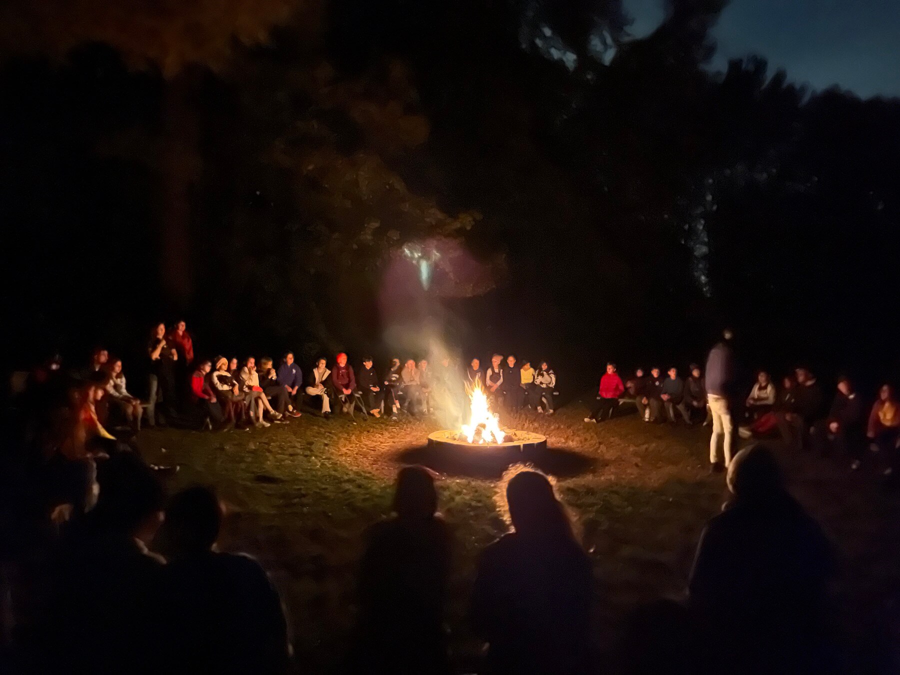 Ethical Culture students, faculty, and staff gather around a bonfire at night.