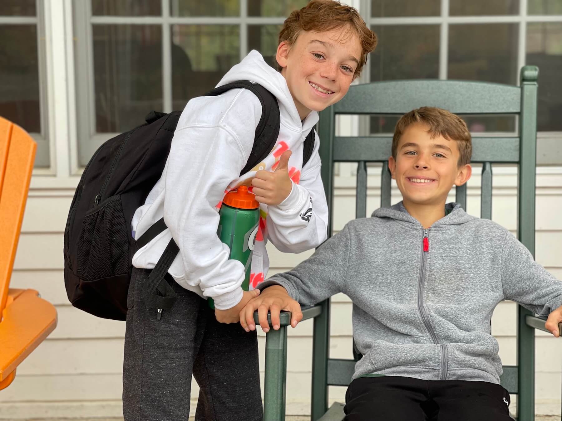 Two Ethical Culture 5th Graders smile at camera on front porch of building. One student sits in a rocking chair, the other is standing.