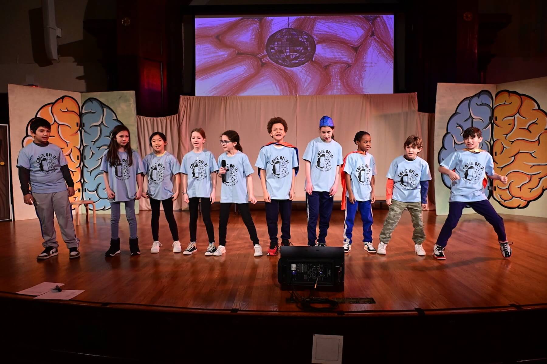 Ethical Culture students dressed in blue t-shirts smile onstage during 4th Grade play performance.