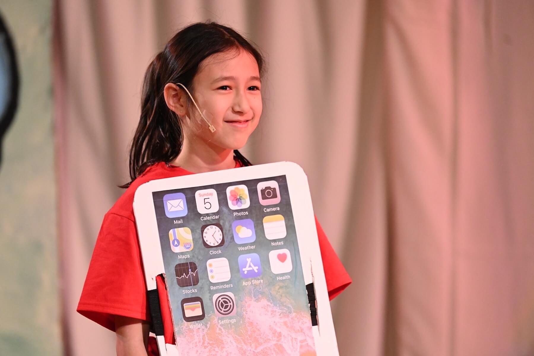 Ethical Culture student dressed as an iPhone smiles onstage during 4th Grade play performance.