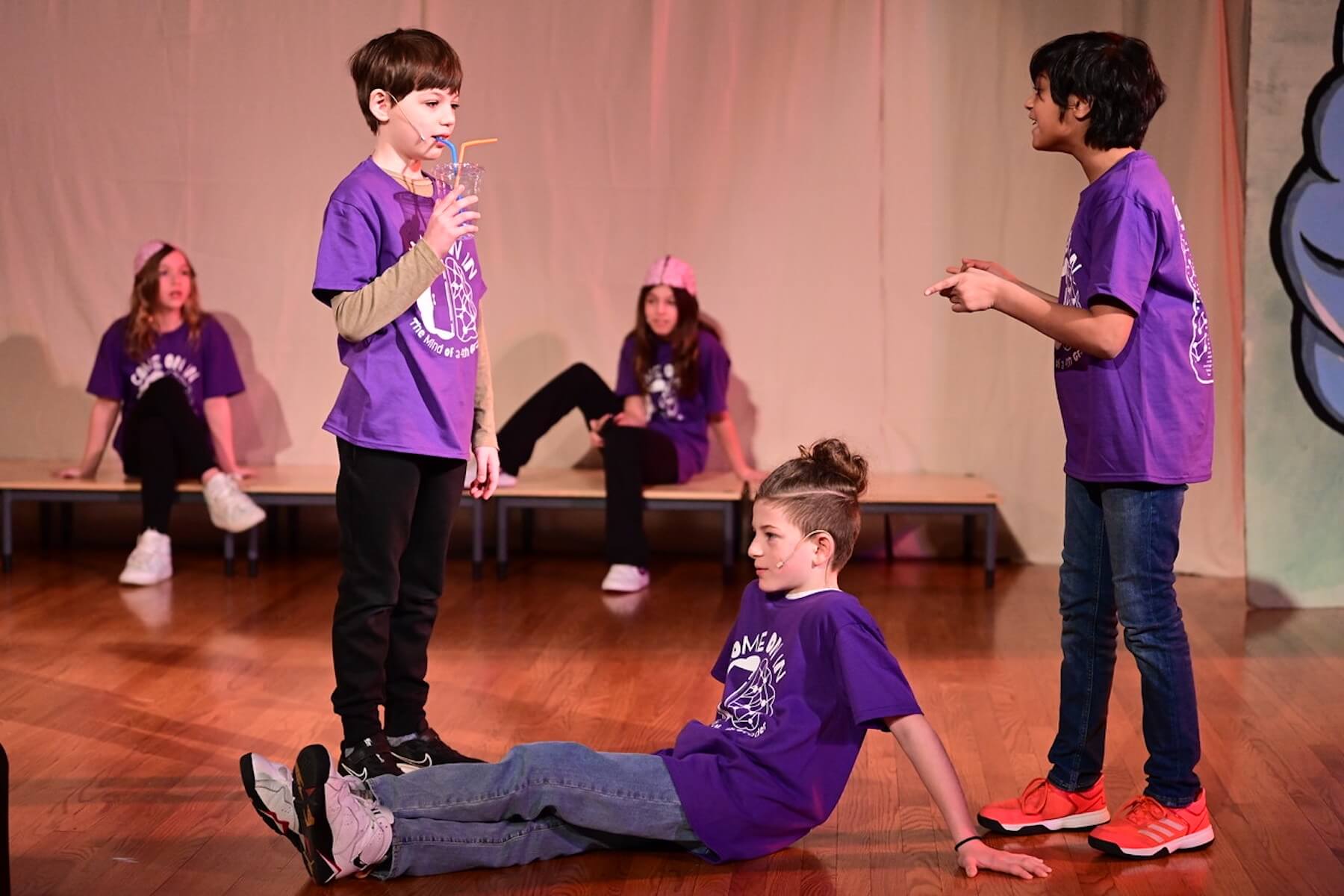 Three Ethical Culture students dressed in purple perform on stage during 4th Grade play. One student sits on stage with legs crossed while two others stand engaged in conversation.