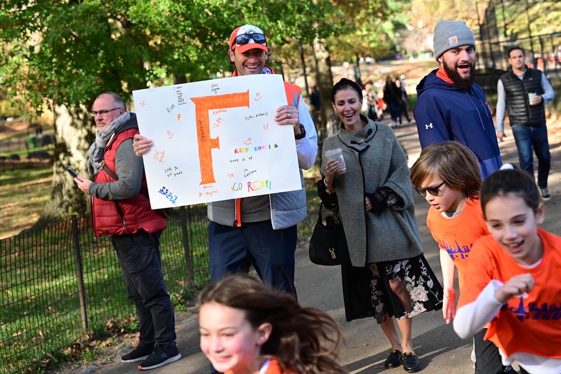 Two ECFS parents stand holding sign that reads has an orange Fieldston "F" on it. Students in the foreground smile and run.