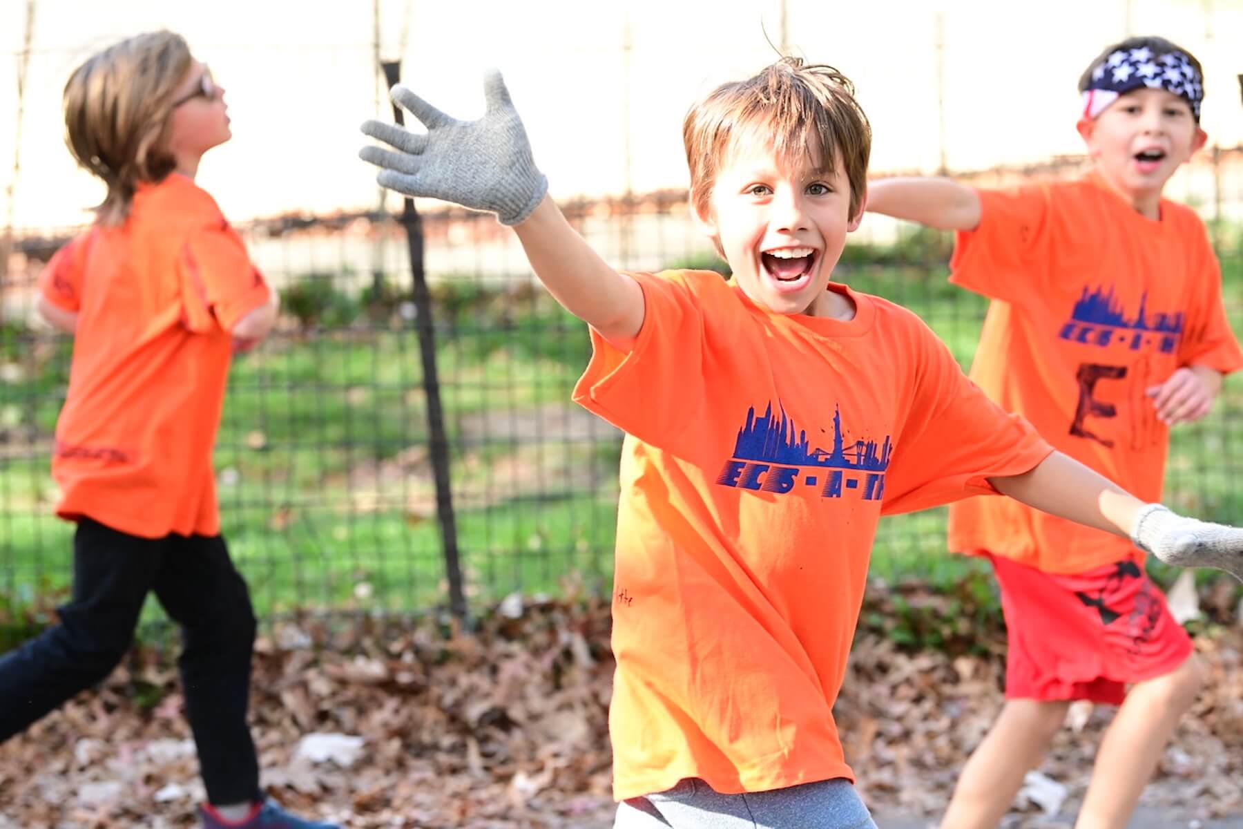 Student wearing orange shirt and mitten smiles and waves to camera as he runs.