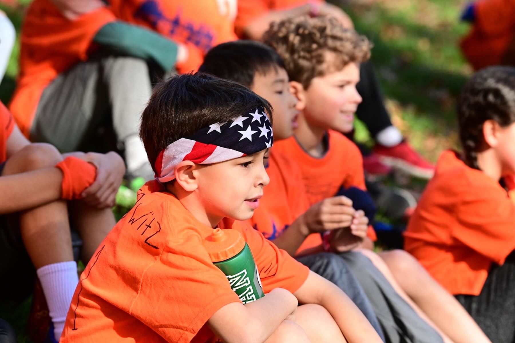 Student sits with his water bottle wearing orange shirt and red, white, and blue bandana.