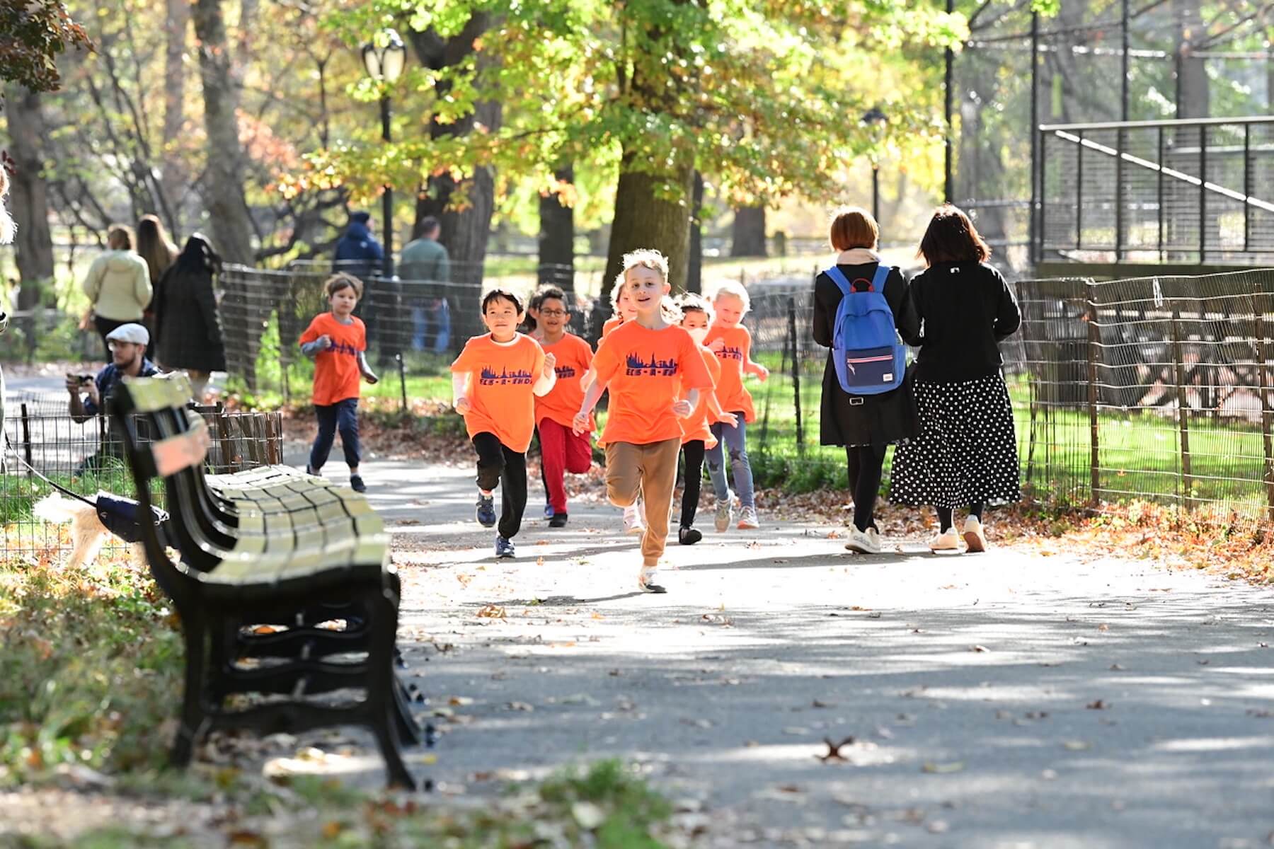 A group of students run together in Central Park wearing orange shirts.