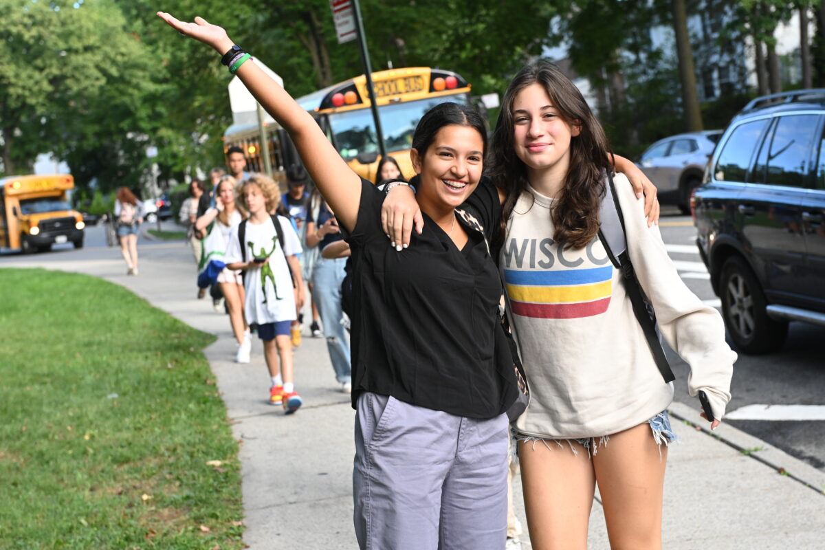 Two students with their arms around each other smile at the camera as they walk from the bus
