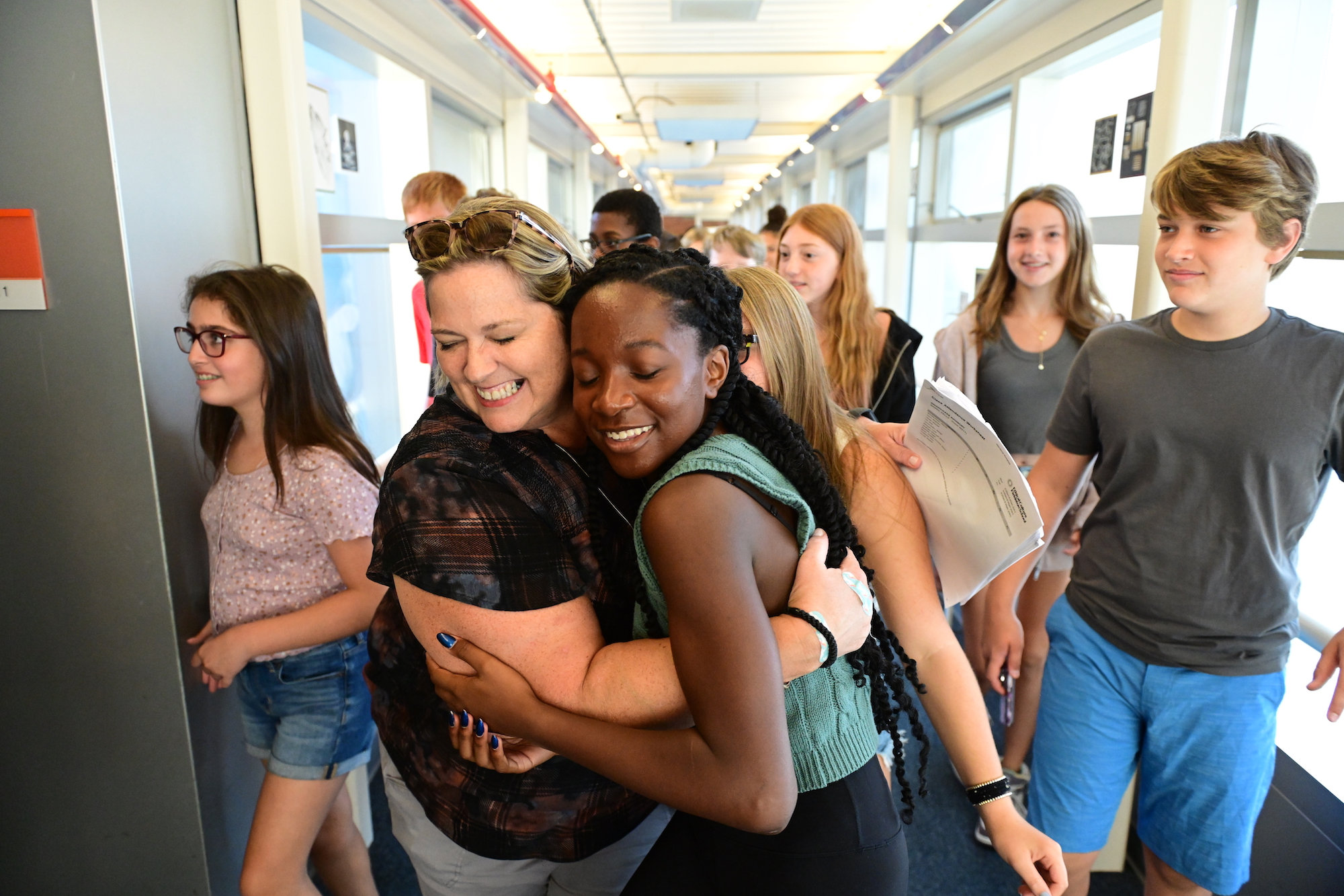 A Fieldston Middle student and a teacher smile and hug on the first day of school as other students walk past them in a hallway