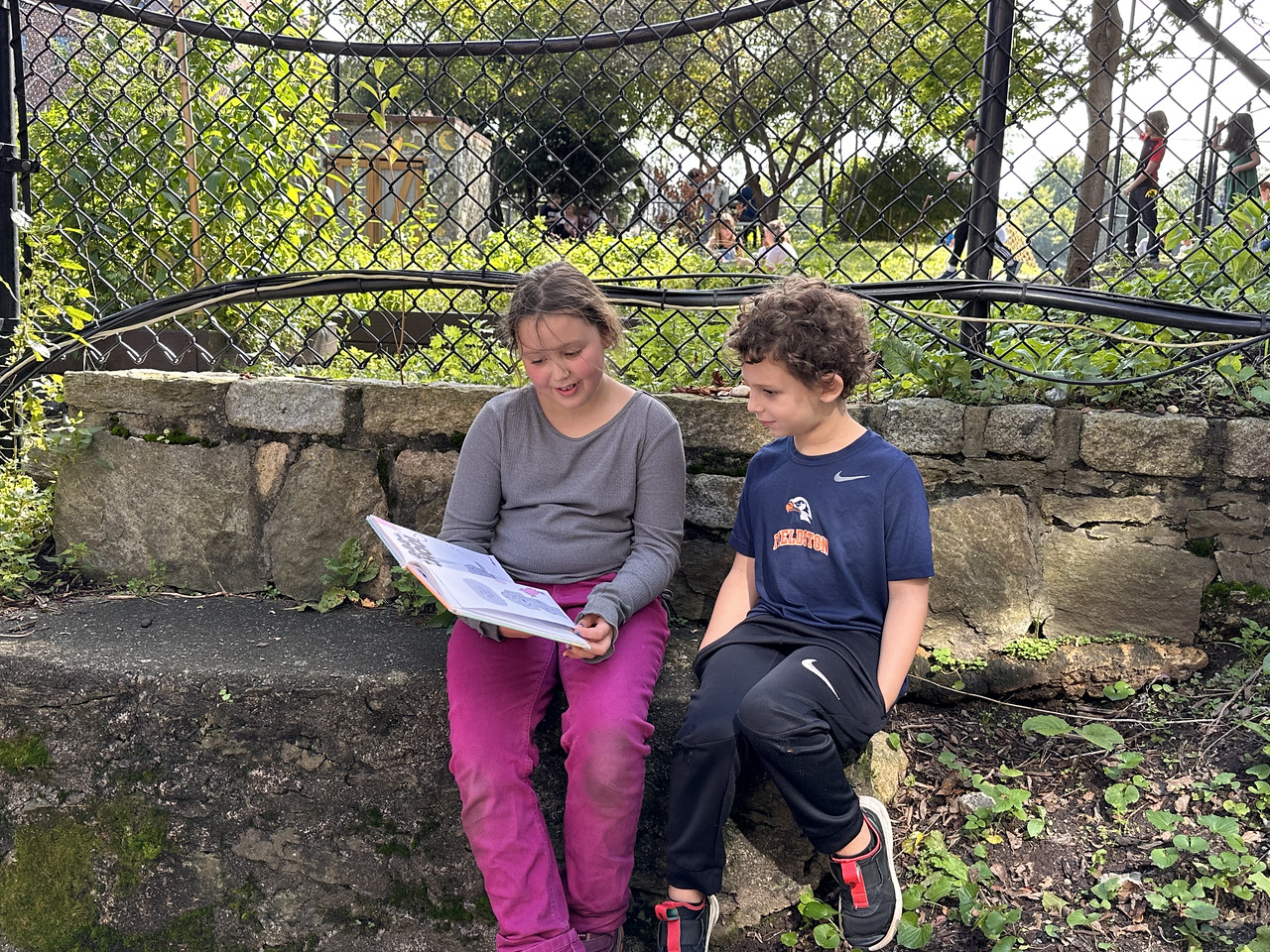 Two Fieldston Lower students read together while sitting on a rock in the outdoor garden.