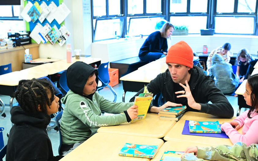 Fieldston Lower student looks intently at his book as teacher explains a part of the text. Other students look on.