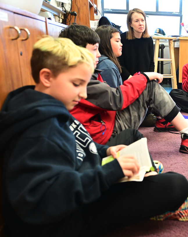 Close-up of Fieldston Lower 4th Grader reading text; other students engage in conversation in the background.