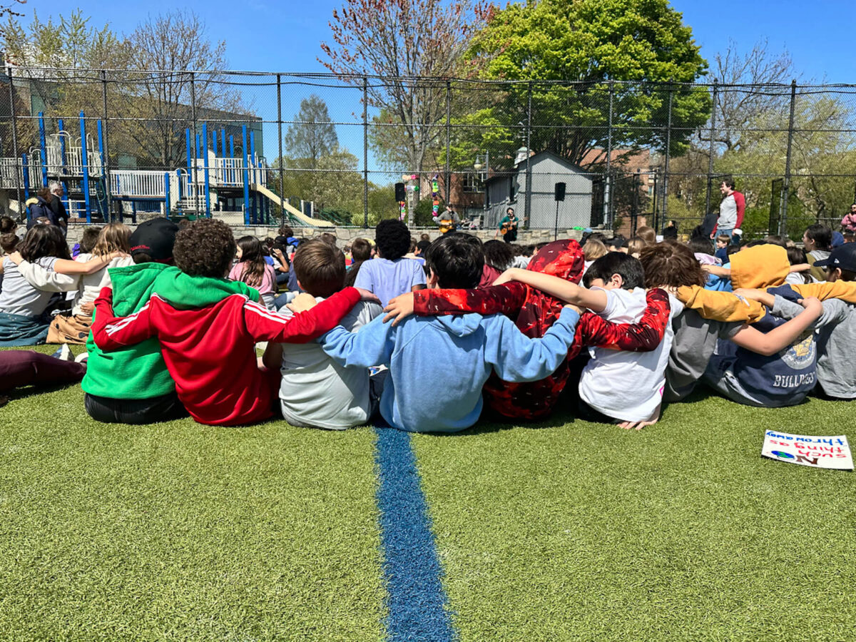 Fieldston Lower 5th Grade students sit on lower field with their arms around one another while they sing the Fieldston Lower School song.