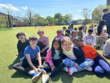 Group of 4th Grade Fieldston Lower students sit together smiling on the lower field at the Earth Day assembly.