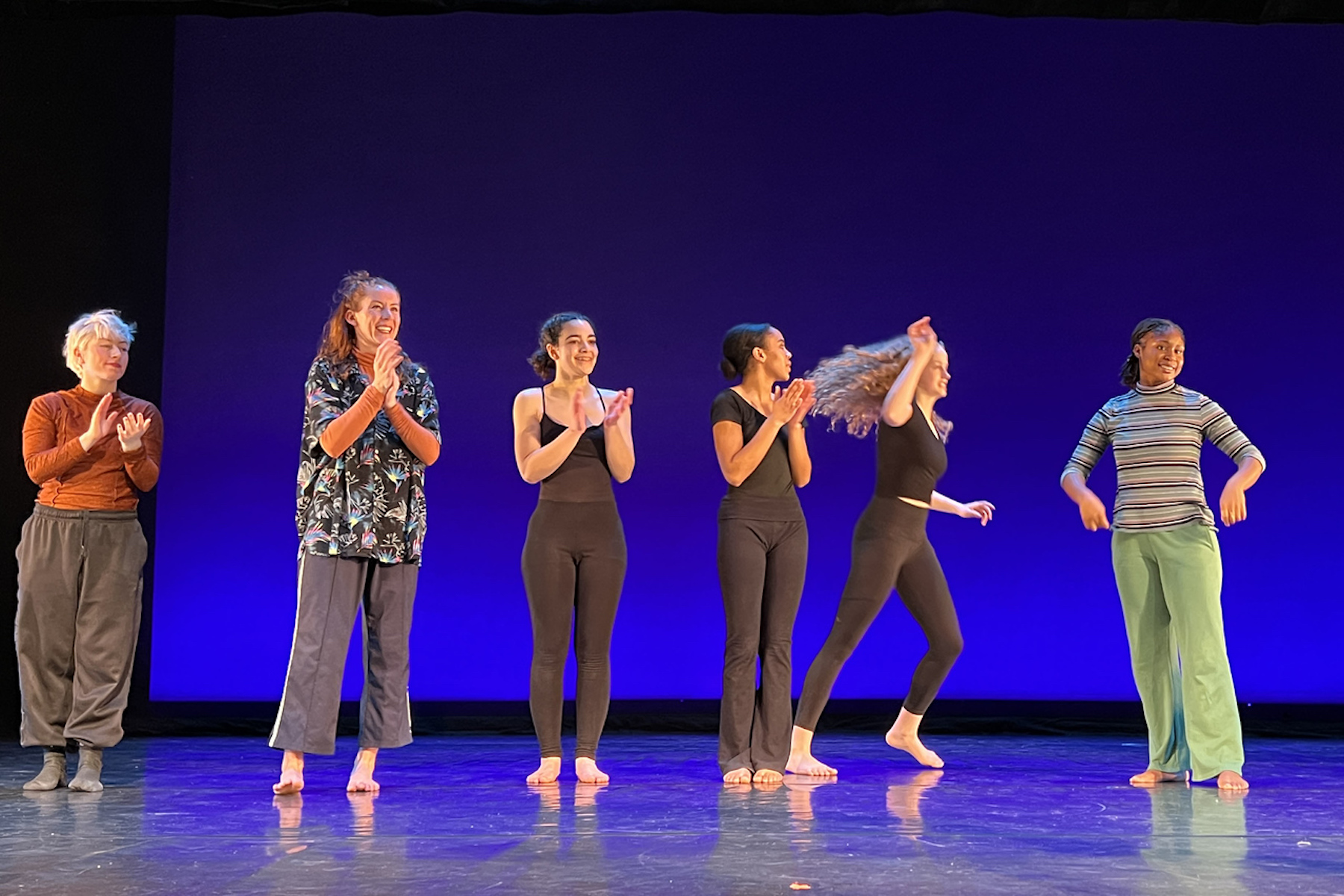 Performers stand in a line on stage, some are clapping, others smiling.