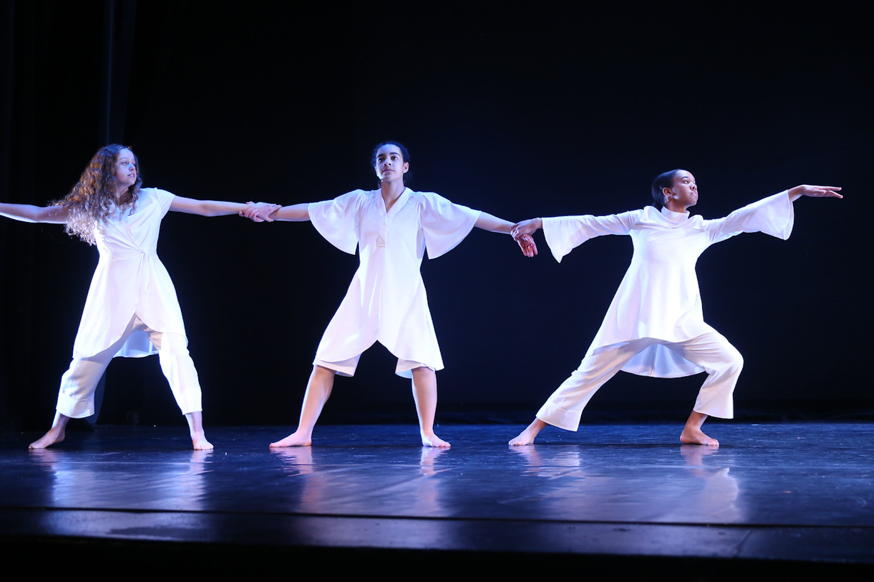 Three Fieldston Upper student dancers perform on stage, wearing all white and holding hands.