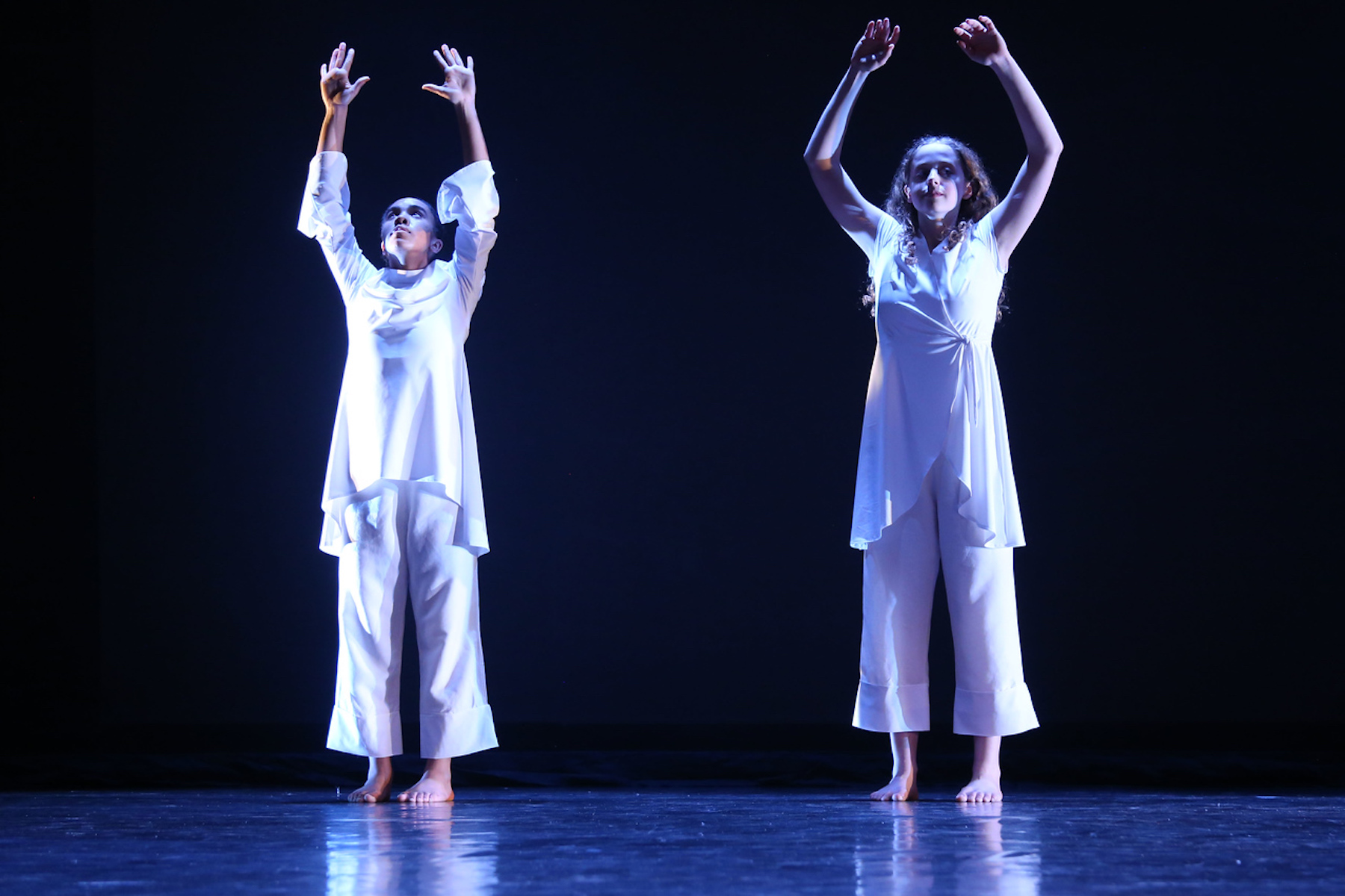 Two Fieldston Upper Dancers perform on stage. They reach their arms up to the ceiling, and wear all white clothing.