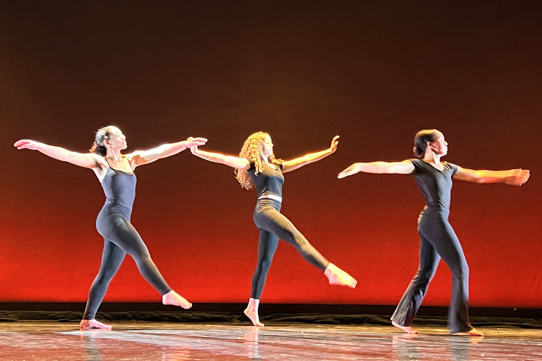 Three Fieldston Upper dancers perform on stage, wearing all black; the stage lighting is a dark red tone.
