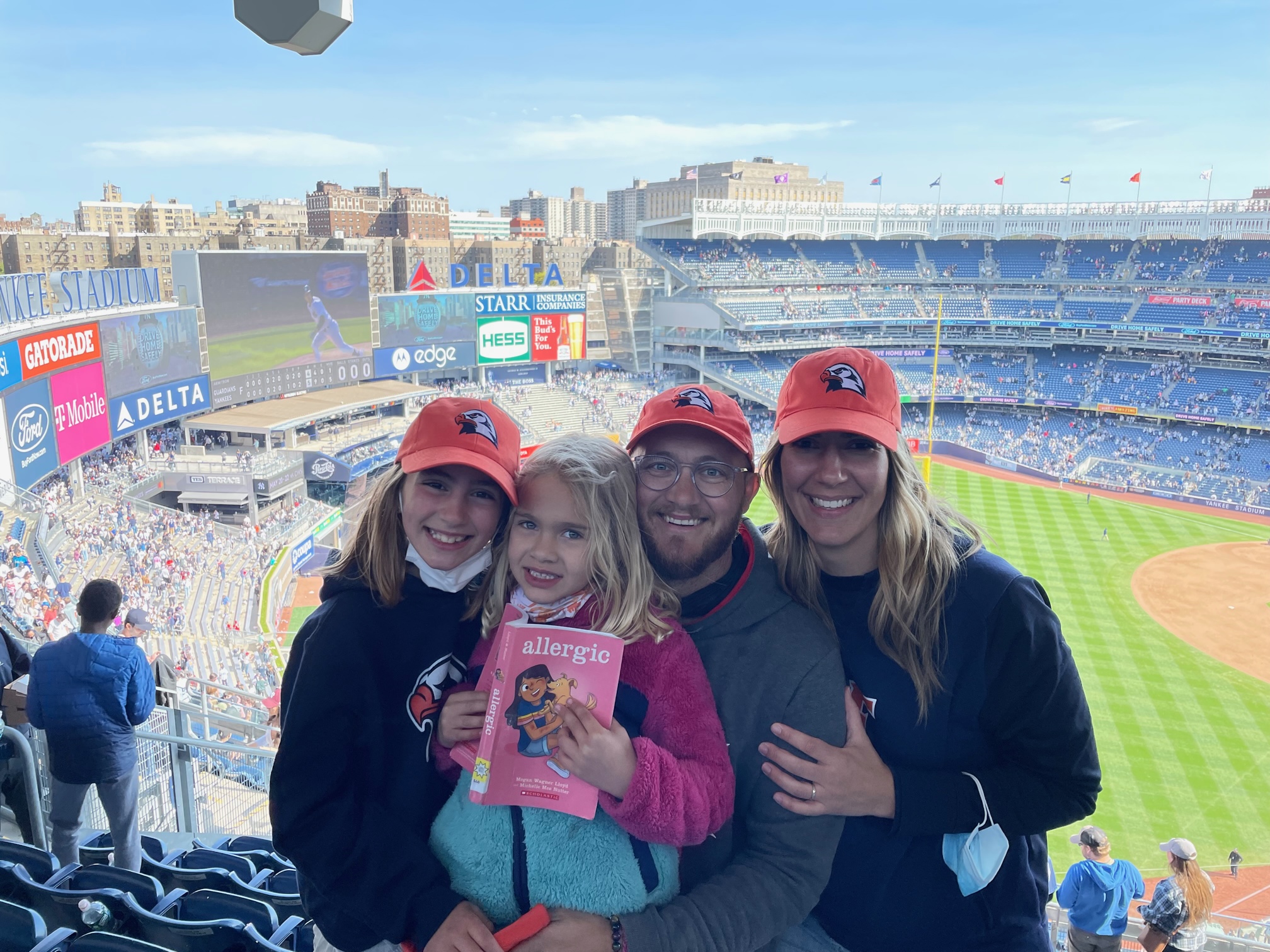 Rose Turshen poses in front of a baseball field with her husband and two children while wearing orange ECFS Eagle hats.