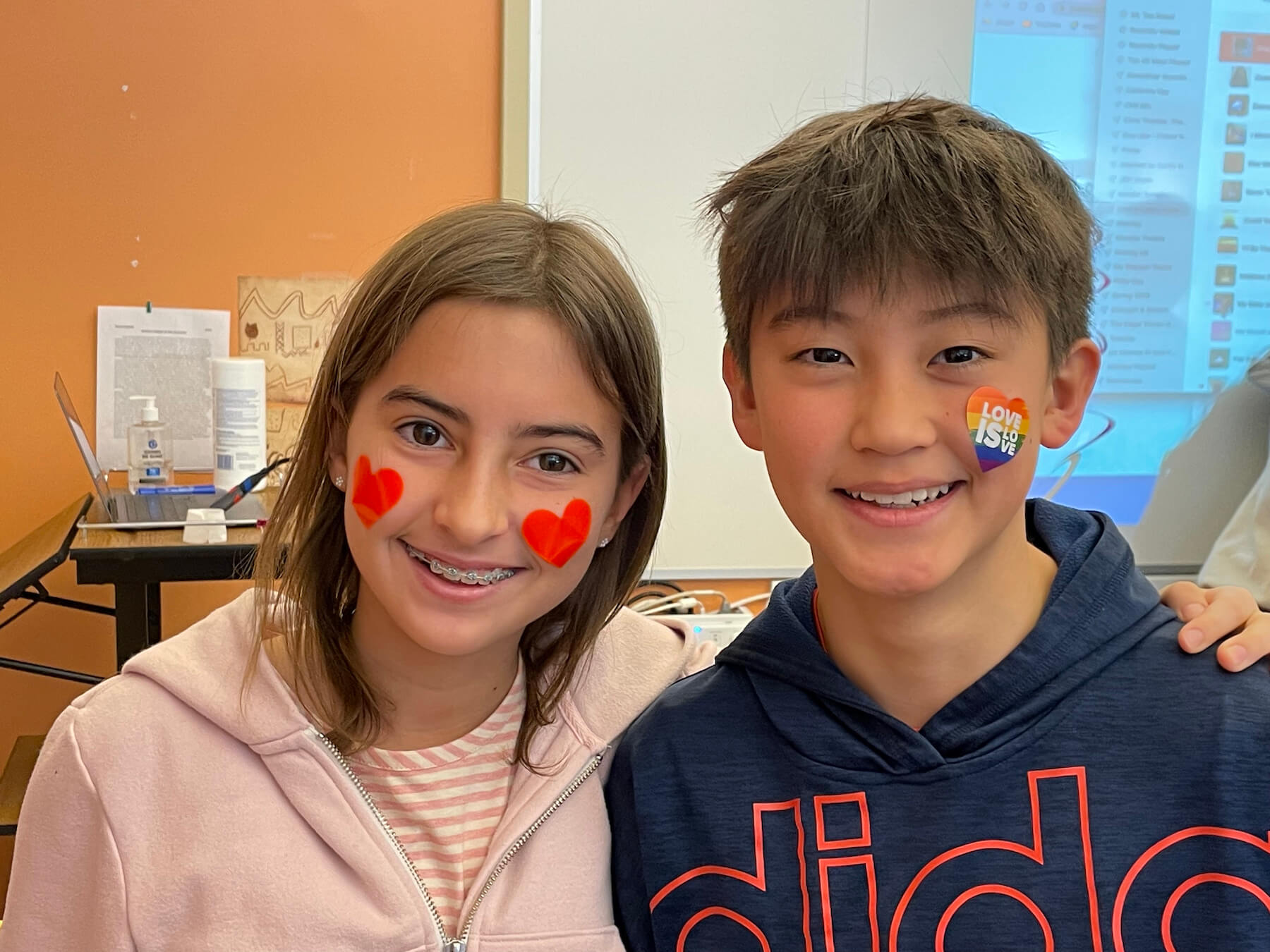 One Fieldston Middle student wearing two heart stickers on her face stands next to a Fieldston Middle student wearing a Love is Love heart sticker on his face.