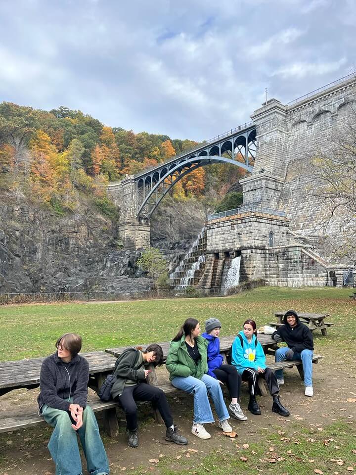 Students sit on a bench outdoors next to a large bridge in a park.