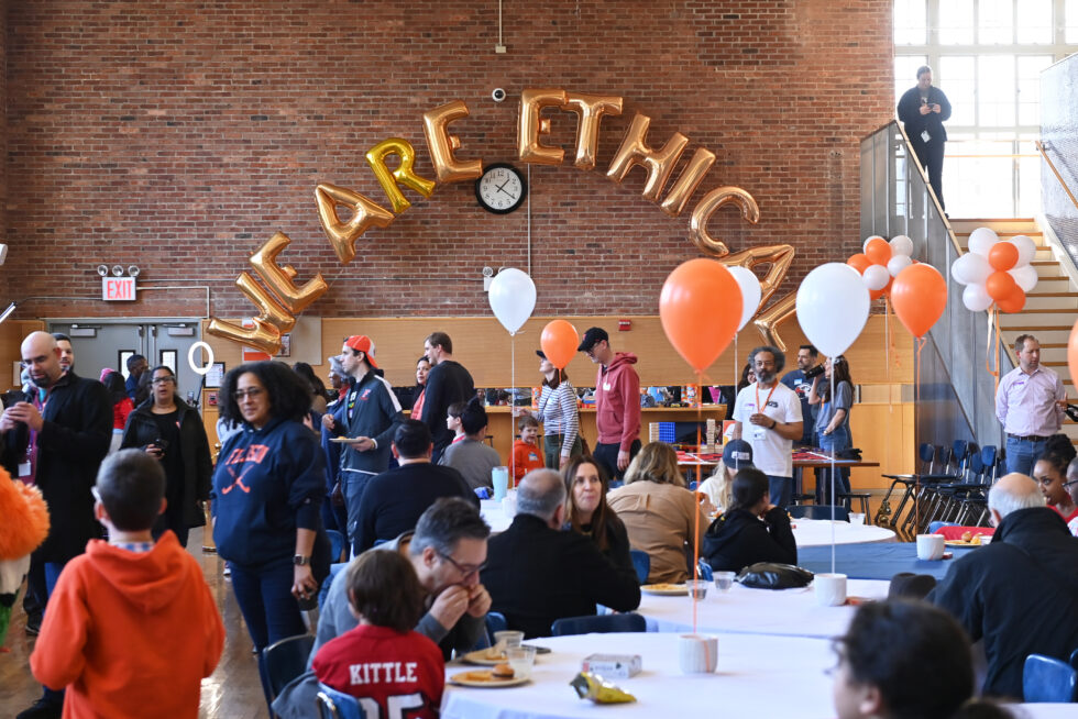 A large group of people sits and stands in the Ethical Culture Fieldston School dining room, surrounded by orange and white balloons and with 