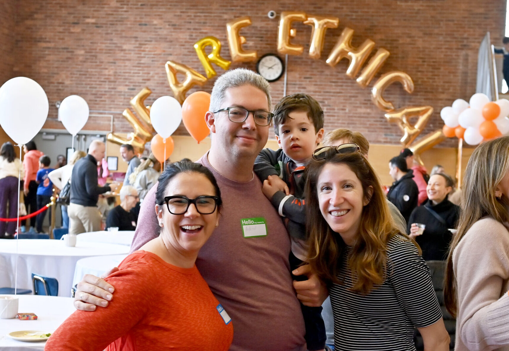 A small group of adults, one holding a young child, pose in front of gold balloons spelling out "We Are Ethical"